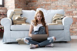 Junge Frau, die auf der Couch mit Laptop und vielen Büchern ruht